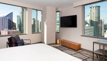 A modern hotel room with large windows showing a cityscape view, a wall-mounted TV, and minimalistic furniture, including a chair and a table.