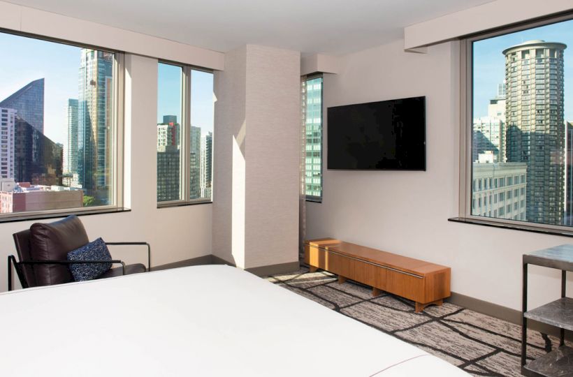 A modern hotel room with large windows showing a cityscape view, a wall-mounted TV, and minimalistic furniture, including a chair and a table.