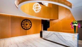 A modern interior lobby with a white marble reception desk, large wall clock, wood paneling, and a contemporary spiral light fixture.