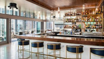 A modern bar with high stools, a wooden counter, and a fully stocked bar in a contemporary setting with large windows overlooking the city.
