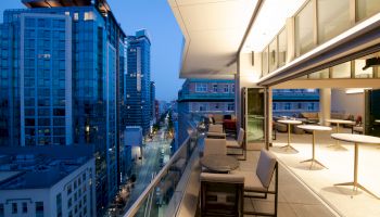 A modern cityscape at dusk featuring tall buildings and a spacious balcony with seating and tables overlooking a street below.