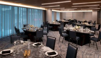 A formal dining room with round tables set for a banquet, adorned with black napkins, glassware, and floral centerpieces, under modern lighting.