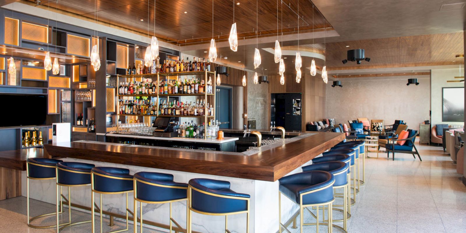 A modern bar area with a well-stocked bar, blue stools, and stylish decor; seating extends to a cozy sitting area in the background.