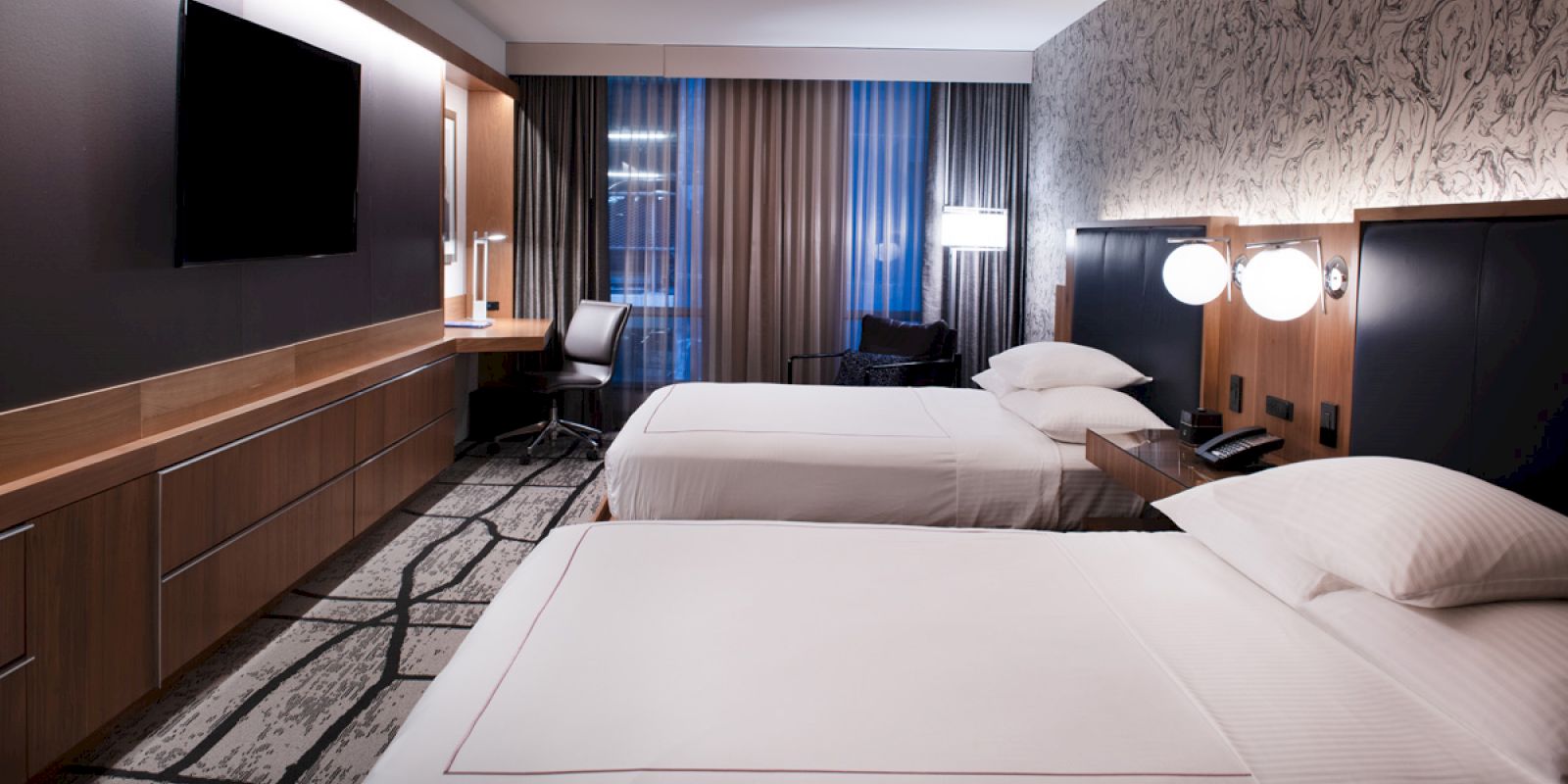 A modern hotel room with two beds, a flat-screen TV, a desk with a chair, and a large window with curtains.