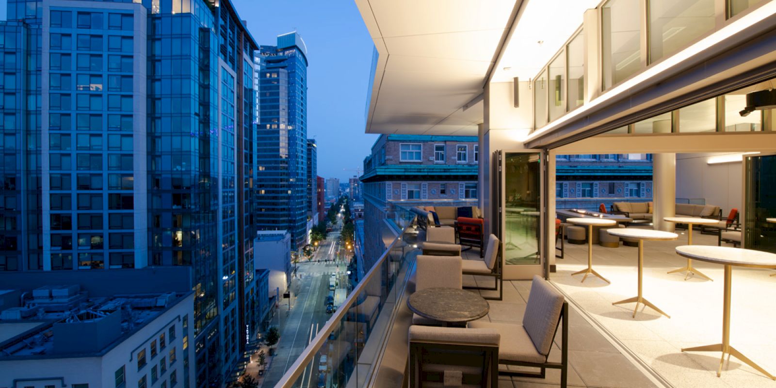 A modern balcony in a cityscape with high-rise buildings, outdoor seating, and tables at dusk. The interior has more seating and ample lighting.