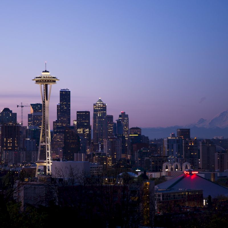 A city skyline at dusk featuring the Space Needle and Mount Rainier in the background under a twilight sky.