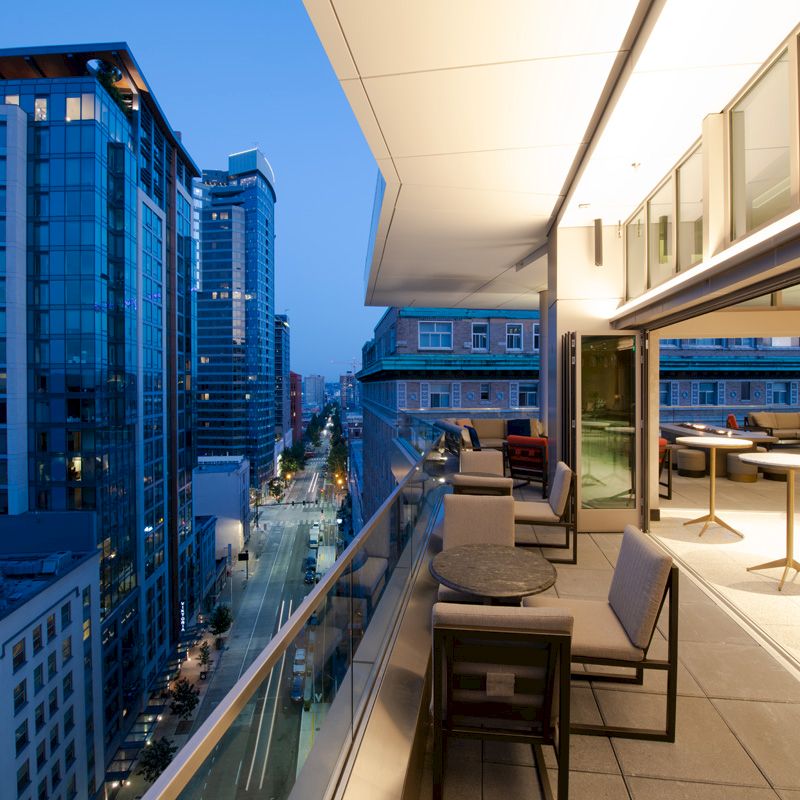 This image shows a modern balcony with seating, overlooking an urban street and tall buildings at dusk. Indoor and outdoor areas are visible.