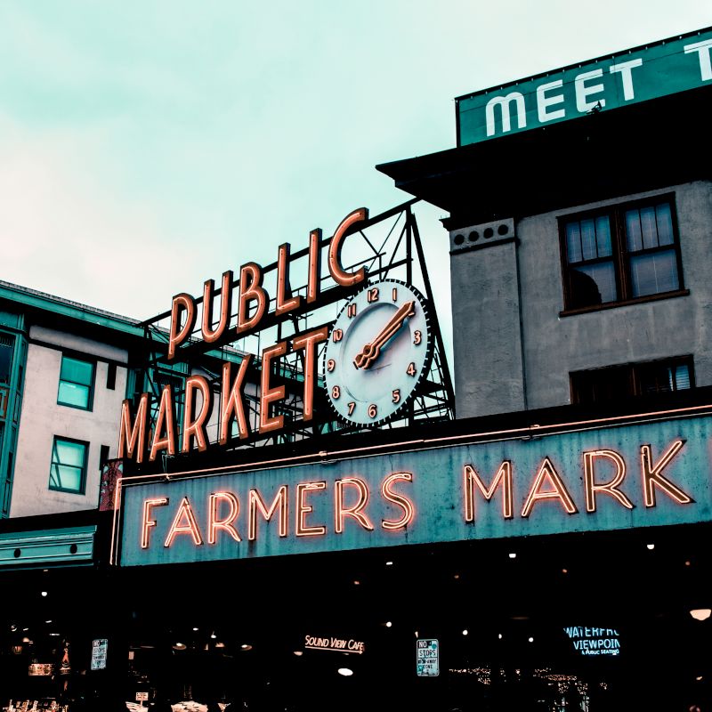 The image shows an iconic "Public Market" sign with a clock, and a lit "Farmers Market" sign below, indicating a popular market location.