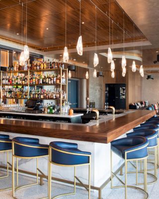 A modern bar with wooden accents, a well-stocked shelf, blue bar stools, hanging lights, and a seating area in the background.