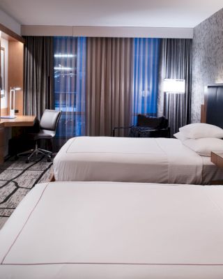 A modern hotel room with two neatly made beds, a wall-mounted TV, a desk, chair, and large windows with curtains.