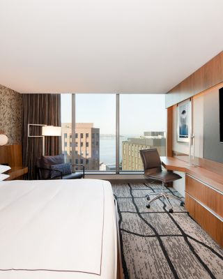 The image shows a modern hotel room with a large bed, a desk with a chair, and a large TV. The room has a window with a city view.
