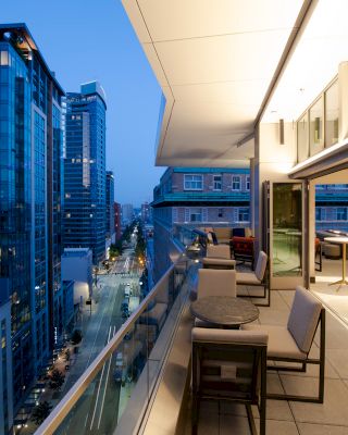 A modern balcony with seating and tables overlooks a city street at dusk, surrounded by tall buildings and illuminated by evening lights.