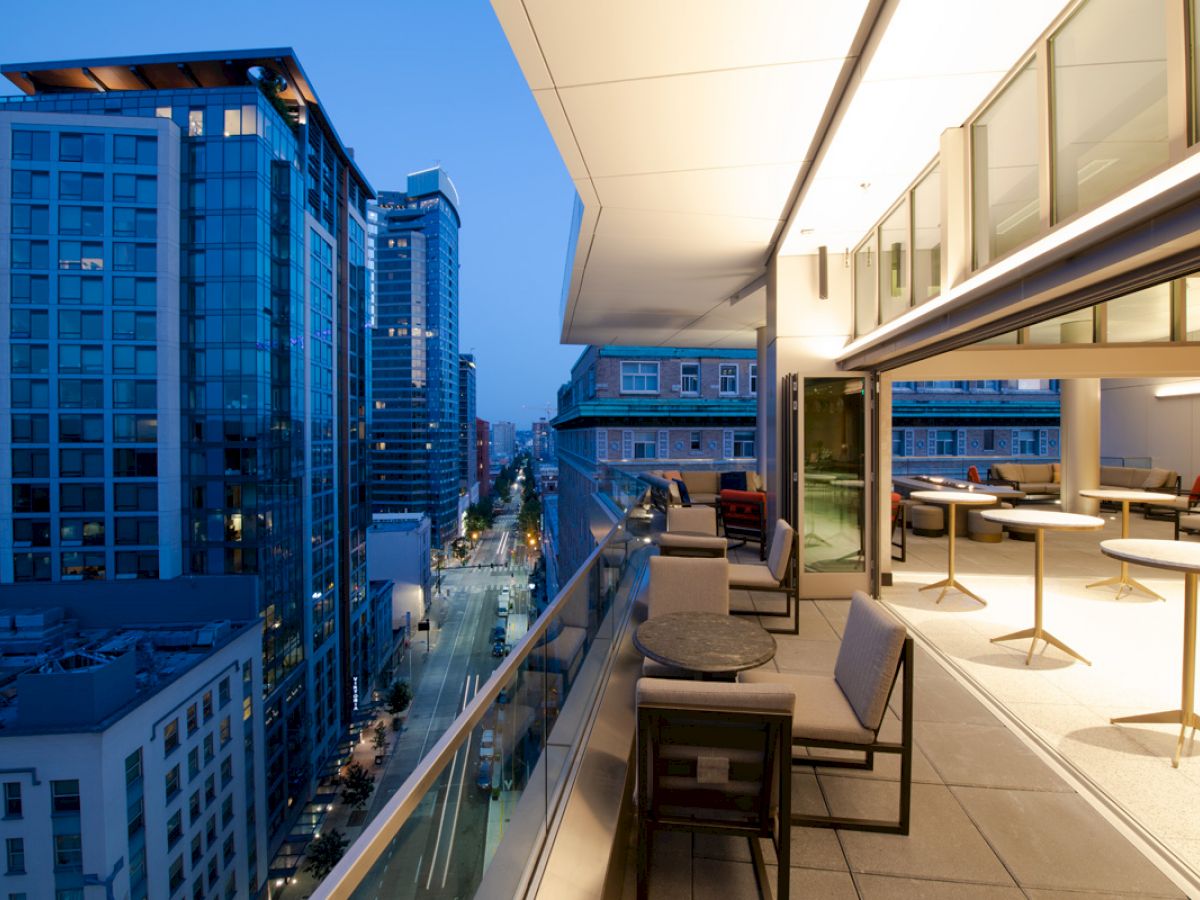 A modern rooftop balcony with seating areas overlooks a bustling cityscape with high-rise buildings during twilight, illuminated by city lights.