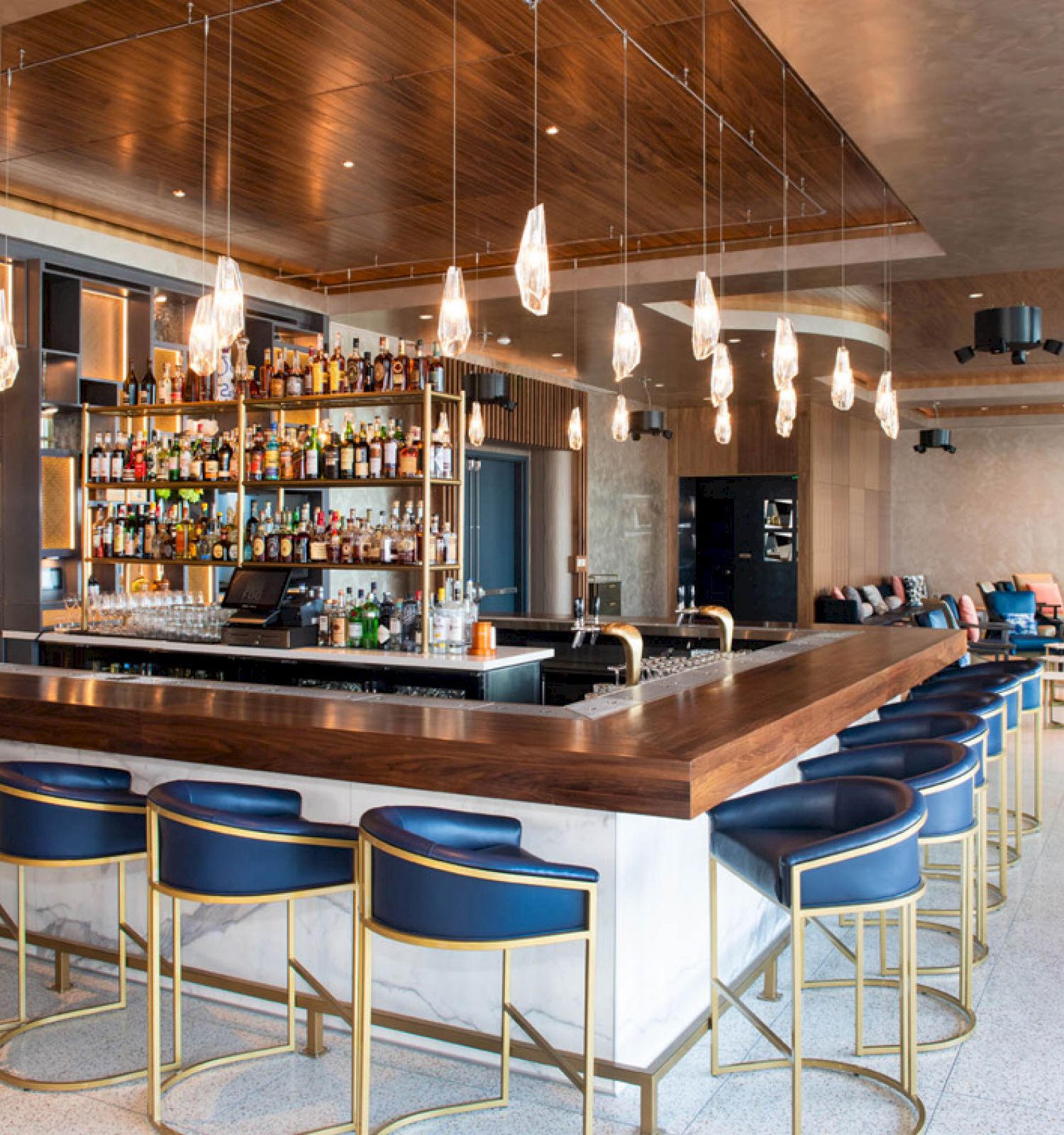 The image shows a modern bar area with blue bar stools, hanging lights, and a fully stocked bar. Seating areas are visible in the background.