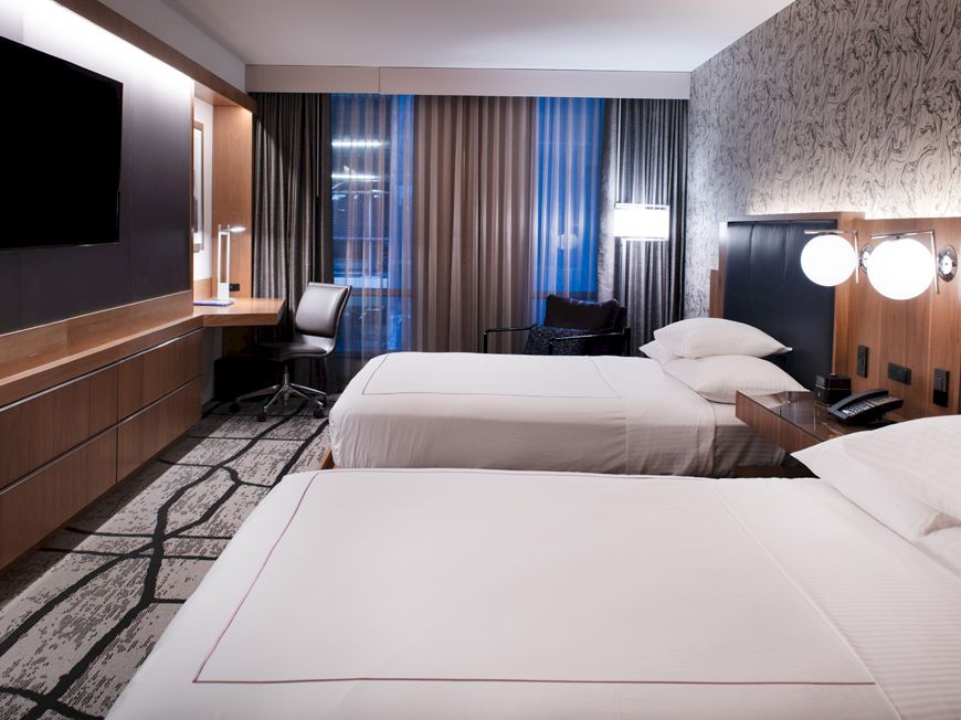 The image shows a modern hotel room with two neatly made beds, a TV, a desk, and large windows with curtains.
