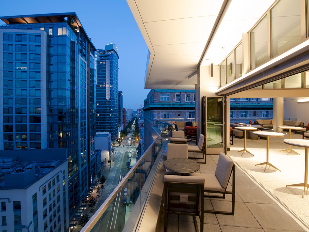 A modern balcony with chairs and tables overlooks a city street with high-rise buildings, illuminated at twilight, providing a scenic urban view.