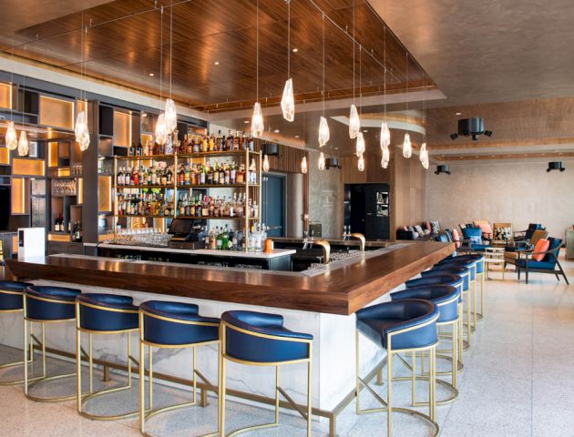 A modern bar with blue barstools, overhead hanging lights, and a well-stocked liquor shelf. There are additional seating areas in the background.