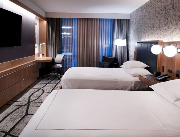 A modern hotel room with two double beds, a flat-screen TV, a desk, a chair, bedside lamps, and curtains covering a window view, is seen.
