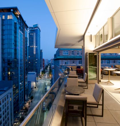 A modern city view at dusk from a rooftop terrace with seating and tables, overlooking a street lined with tall buildings.