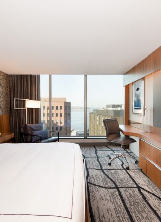 A modern hotel room with a large bed, wall-mounted TV, desk, armchair, and floor-to-ceiling window with a city view outside.