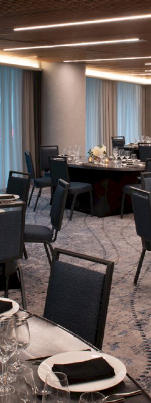 The image shows an elegant dining room with round tables set up for an event, featuring black chairs, plates, glasses, and floral centerpieces.