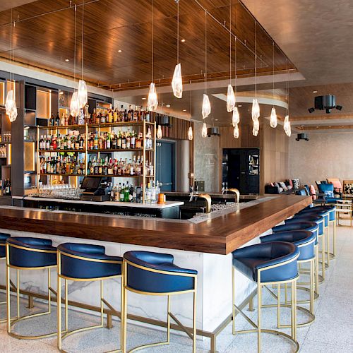 A modern bar setup with blue and gold bar stools, a well-stocked shelf of liquor behind the bar, and contemporary light fixtures.