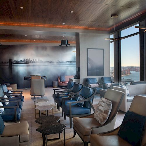 A modern lounge area with comfortable seating, large windows revealing a scenic view of a waterfront and a Ferris wheel.