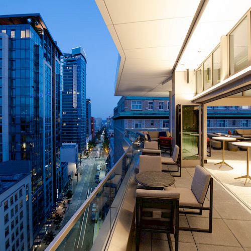The image shows a modern cityscape during dusk with tall buildings, a rooftop terrace with tables and chairs, and a view of the street below.