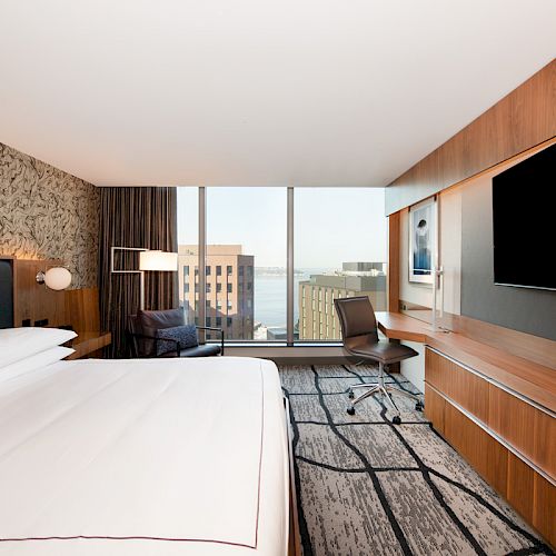 The image shows a modern hotel room with a large bed, a desk with a chair, a flat-screen TV, and a window with a cityscape view.