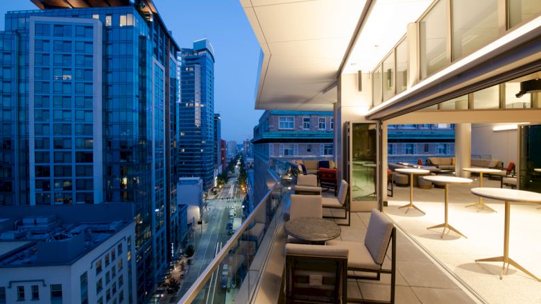 An outdoor terrace with seating and tables overlooking a cityscape with high-rise buildings at dusk, featuring a modern and urban setting.