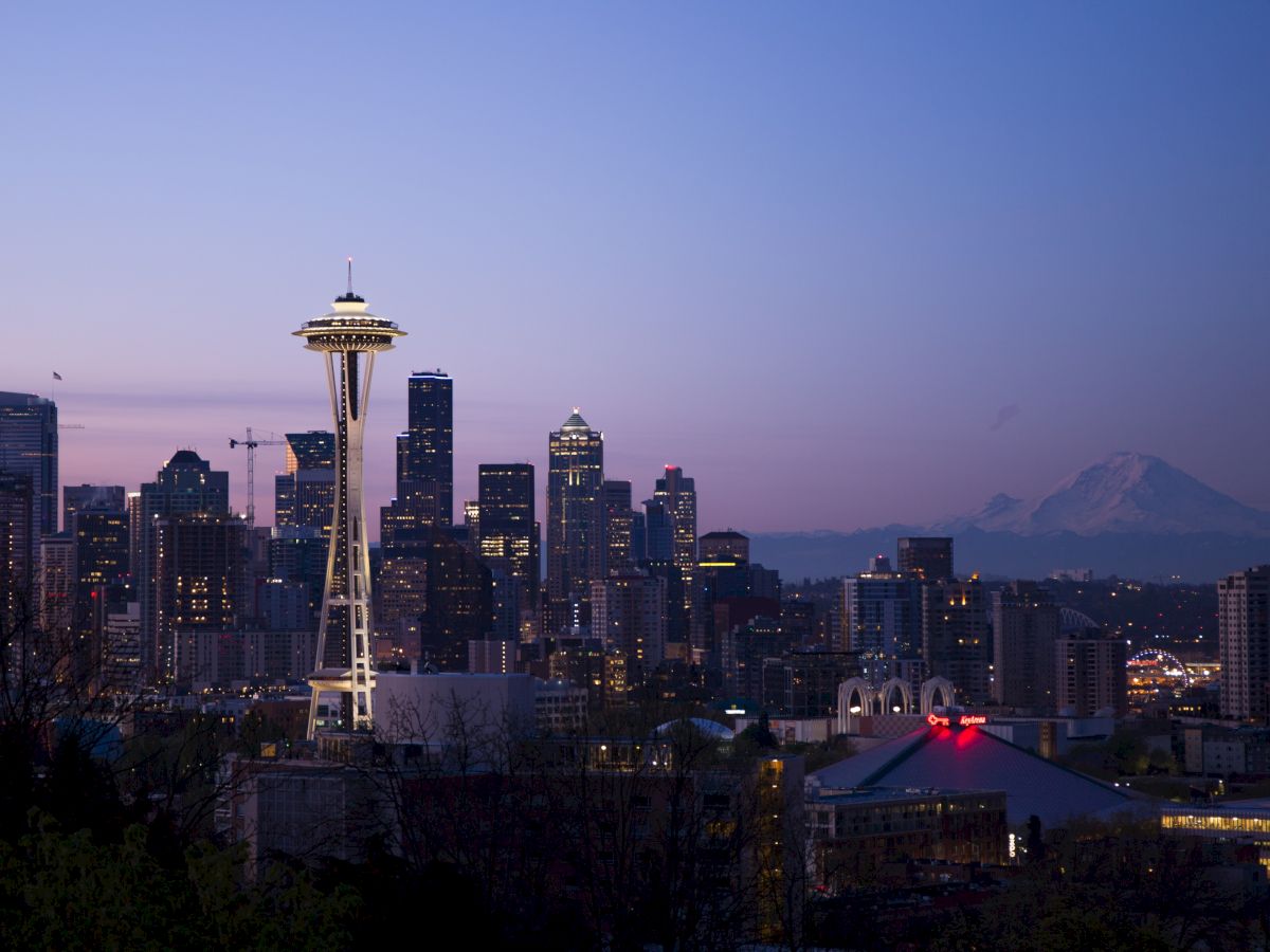 A skyline of Seattle at dusk, featuring the Space Needle with Mount Rainier in the background and several buildings illuminated by lights.