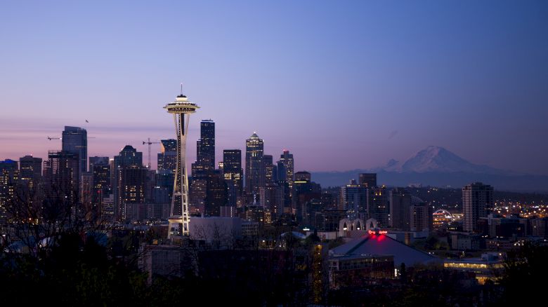 A skyline of Seattle at dusk, featuring the Space Needle with Mount Rainier in the background and several buildings illuminated by lights.
