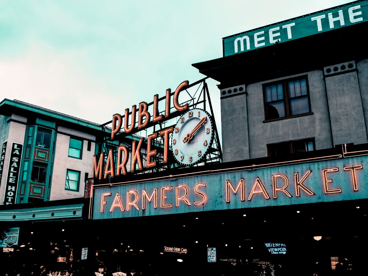 A neon sign reading "Public Market" and "Farmers Market" with a clock is displayed on the exterior of a building. Other signs and structures are visible.