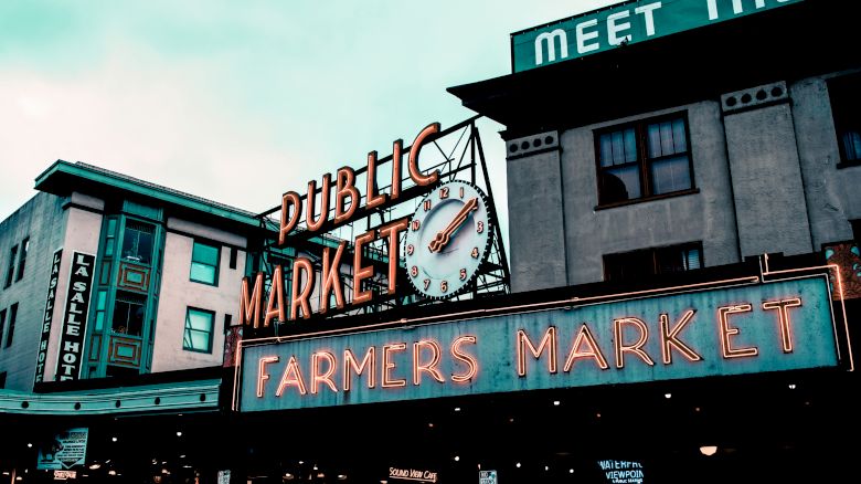 A neon sign reading "Public Market" and "Farmers Market" with a clock is displayed on the exterior of a building. Other signs and structures are visible.