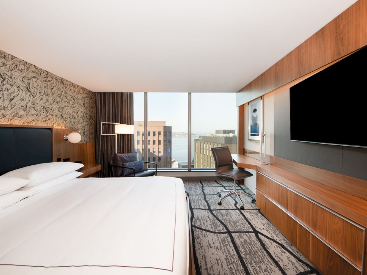 This image shows a modern hotel room with a large bed, a desk with a chair, a large TV, and a window offering a cityscape view.