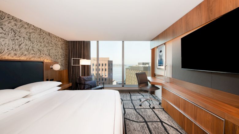 This image shows a modern hotel room with a large bed, a desk with a chair, a large TV, and a window offering a cityscape view.