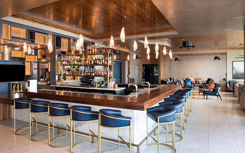 A stylish bar with a wooden countertop, blue bar stools, pendant lights, and a fully stocked back bar. Seating areas are visible in the background.