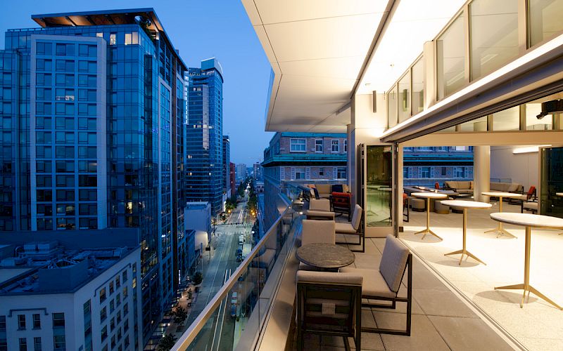 A modern balcony view featuring outdoor seating, city streets, and high-rise buildings, captured during the evening with glowing lights.