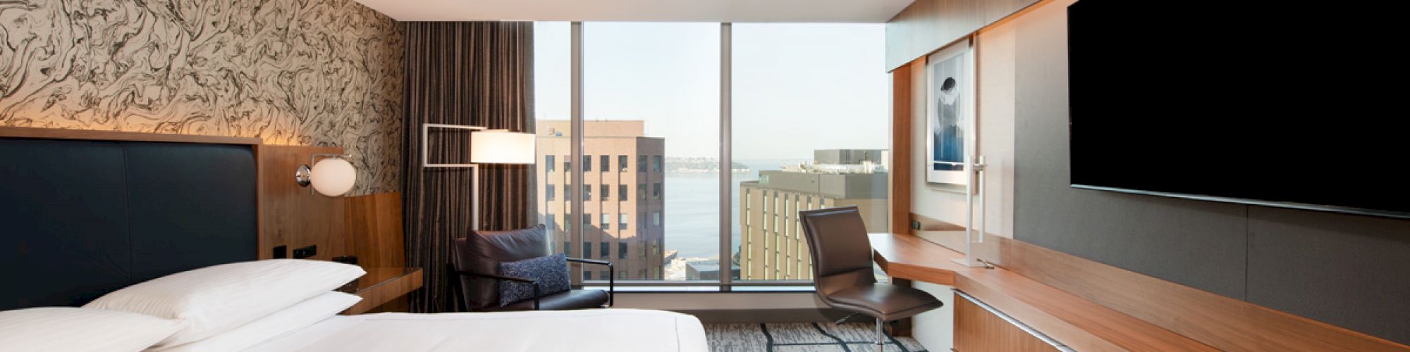 A modern hotel room with a large bed, desk, chair, flat-screen TV, and a window offering a city and waterfront view.