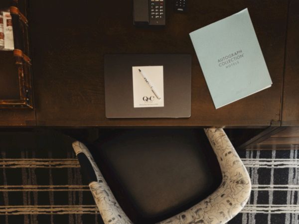 A desk with a chair, a TV remote, a blue card, and a small white card on a dark surface, plus a plaid rug beneath the chair, neat and tidy.