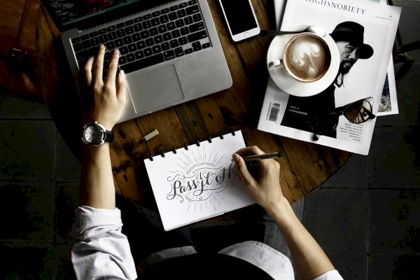 An overhead view of a person working on a laptop at a wooden table, with a smartphone, coffee, magazines, and a notepad with handwritten text.