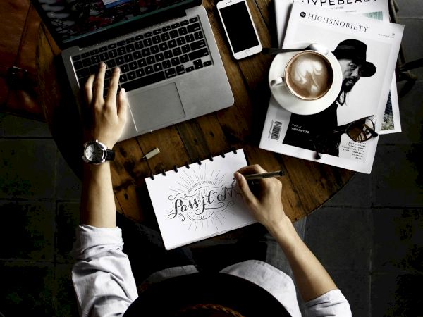 An overhead view of a person working on a laptop at a wooden table, with a smartphone, coffee, magazines, and a notepad with handwritten text.